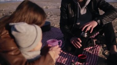 Genç ve şık bir aile, günlük kıyafetleriyle kıyı kenarında oturmuş piknikte sıcak bir şeyler içiyorlar. Anne oğluna bir fincan çay verir. Soğuk hava, hafta sonu tatili.