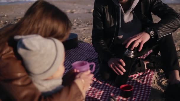 Joven y elegante familia en ropa casual sentada a cuadros junto al agua y tomando bebidas calientes en el picnic. Mamá le da una taza de té a su hijo. Clima frío, recreación de fin de semana — Vídeo de stock