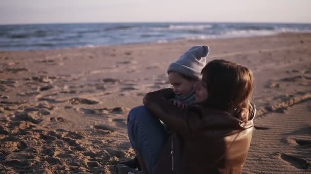 Ευτυχισμένη μητέρα αγκαλιάζει το παιδί της στην παραλία του φθινοπώρου. Χαριτωμένο παιδί αγκαλιάζει τη μαμά του και πέφτουν στην άμμο. Concept μαμά με ένα παιδί σε εξωτερικούς χώρους, ευτυχισμένη οικογένεια, χαρούμενη παιδική ηλικία, μικρό μωρό — Αρχείο Βίντεο