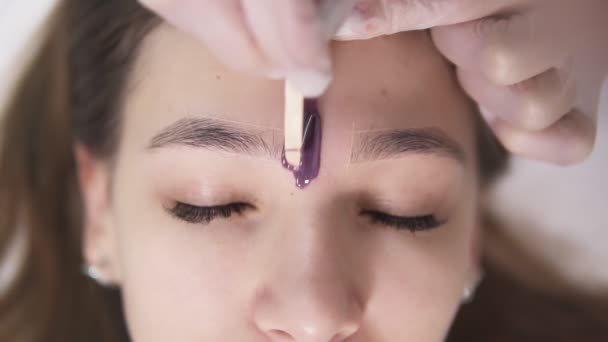 El cliente está acostado en el sofá durante el tratamiento de las cejas en la belleza del estudio, esteticista depilando y moldeando las cejas en el salón de belleza, aplicando un poco de cera entre las cejas. Sobre las imágenes de la cabeza. Despacio. — Vídeo de stock