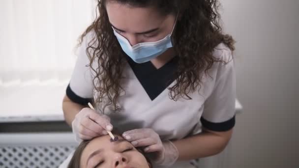 Menina bonita jovem está deitada no sofá durante o tratamento eyebrowes na beleza do estúdio, esteticista depilação e moldar as sobrancelhas no salão de beleza. Mestre aplicando uma linha de cera acima da testa usando um pau — Vídeo de Stock