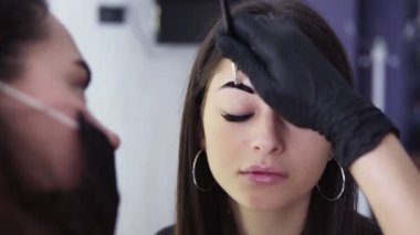 Omuzdan alınan görüntülerde, kafkas güzellik uzmanı kahverengi boya ile genç kadının kaşlarında koyu boya, bir güzellik stüdyosunda boyama işlemi, yakın çekim, ön görünüm