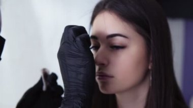 Siyah eldiven bir güzellik beyaz temiz pamuk disk ank küçük kaş fırça kullanarak kaşboya kaldırmak için bir kadın güzellik salonunda bir ziyaret sırasında sandalyede oturur. Yavaş çekim