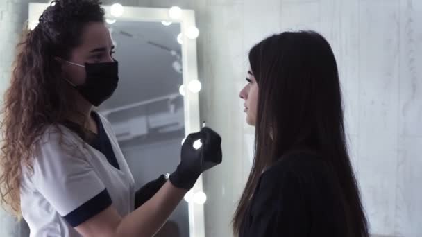 Capelli lunghi estetista caucasica sta applicando vernice scura sulle sopracciglia della giovane donna da tintura marrone, procedura di colorazione in uno studio di bellezza, primo piano, vista laterale — Video Stock