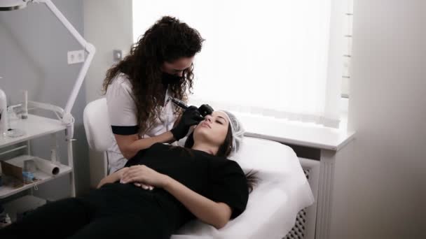 Beautician is drawing the shape or making correction of eyebrows with needle for a woman in a beauty salon. Professional equipment for permanent makeup. Modern cosmetology — Stock Video