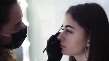Siyah maskeli uzun saçlı beyaz güzellik uzmanı kahverengi boya ile genç kadının kaşlarında koyu boya, güzellik stüdyosunda boyama işlemi, yakın çekim, yan görünüm