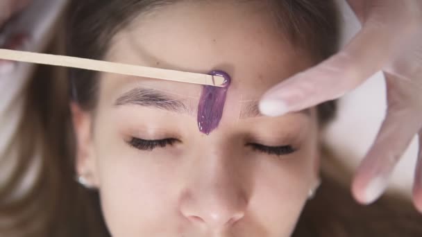 Młoda, kaukaski dziewczyna leży na kanapie podczas leczenia eyebrowes w Studio urody, kosmetyczki depilacji i kształtowania brwi w salonie piękności, usuwając wosk między brwi za pomocą kija. Materiału — Wideo stockowe