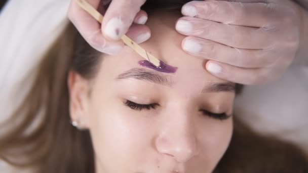 Joven, chica caucásica está acostado en el sofá durante el tratamiento de las cejas en el estudio de belleza, esteticista depilación y dar forma a las cejas en el salón de belleza. Maestro aplicando una línea de cera sobre la frente usando un palo — Vídeo de stock