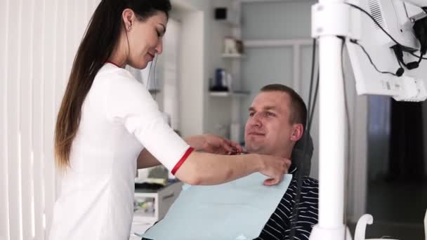 Il dentista femminile dai capelli lunghi prepara il paziente maschile per il trattamento dei denti sullo sfondo dell'armadio stomatologico. Il medico mette un pannolino di copertura sul petto di un giovane su una sedia da dentista — Video Stock