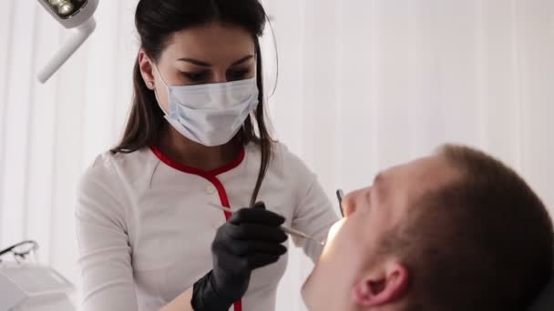 マスクの女性歯科医は患者の口腔を調べる。男性は歯科用椅子、歯科治療の概念に座っています。治療プロセス中にラボコートとマスクの若い歯科医。クローズアップ — ストック動画