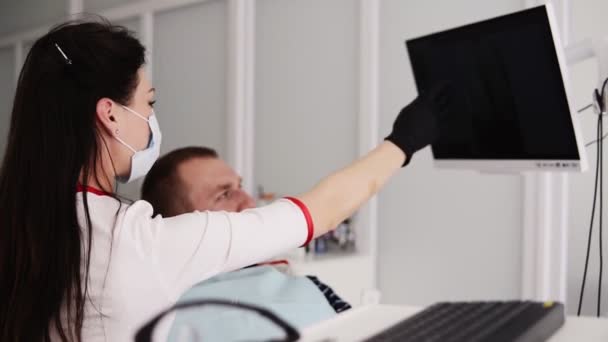 Dentista caucasica in vestaglia bianca, maschera e guanti neri che spiega l'immagine a raggi X sullo schermo nero al paziente maschio. Mostra l'immagine di dente malsano a un cliente. Filmati dal lato — Video Stock