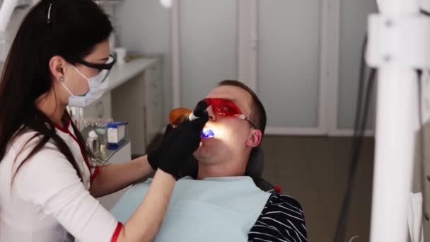 Médica e paciente do sexo masculino em óculos laranja de proteção no moderno ocabinete dental. Dentista está fazendo polimerização de recheios com uma tela protetora laranja — Vídeo de Stock