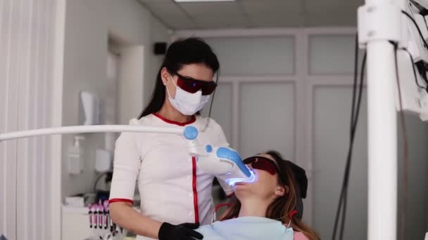 Procedimiento de blanqueamiento dental en clínica odontológica. Mujer joven con gafas protectoras sentada en la silla del dentista. Doctor sosteniendo dispositivo moderno, lámpara ultravioleta con luz azul. Dentista femenina en blanco — Vídeo de stock