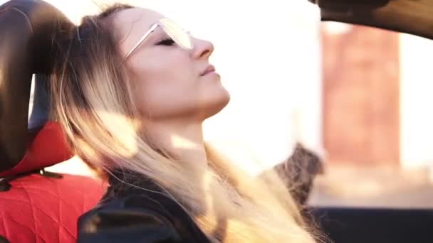 Chica despreocupada y de pelo largo en gafas de sol transparentes que conducen su coche cabriolet en la ciudad. Joven mujer reflexiva disfrutando de las bengalas de las lentes de verano, extienda su mano apoyada en la puerta del automóvil — Vídeo de stock