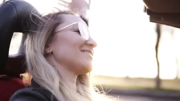 Chica positiva, sonriente y de pelo largo en gafas de sol transparentes que conducen su coche cabriolet en la ciudad. Joven mujer disfrutando conducir, extender su mano apoyada en la puerta del coche - atrapando el viento, cantando — Vídeo de stock