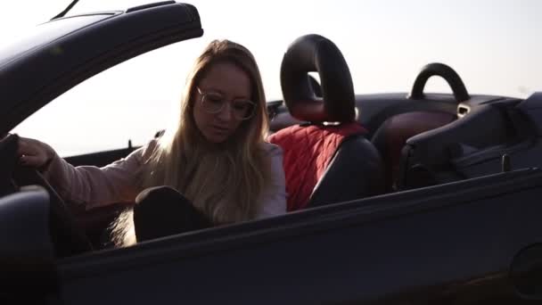 Young woman driver in beige leather jacket with long hair and stylish sunglasses opening the door of her cabriolet car at a stop during a trip to look around. Foggy, blurred background — Stock Video