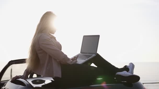 Мандрівник використовує ноутбук у дорожній поїздці. Молода жінка читає і друкує на комп'ютері, використовуючи мобільний Інтернет, розслабляючись на своєму кабріолетному автомобілі з сонячним світлом на фоні — стокове відео