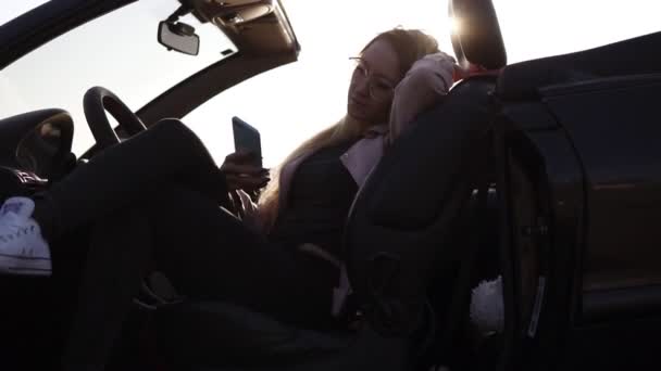 Jonge aantrekkelijke meisje zittend, ontspannen in haar zwarte sport auto en het houden van mobiele telefoon. Ze draagt mosern zonnebrillen en beige jasje. Milde zomerzon op de achtergrond. Lage hoek beelden — Stockvideo