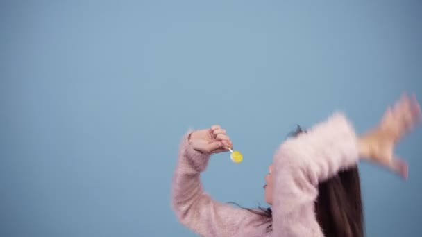 Смешная девушка в розовом свитере веселится, танцует, прыгает и поет под желтый леденец. Портрет красивой молодой девушки, поющей под леденец на синем фоне. Девушка поет в конфетах, как — стоковое видео