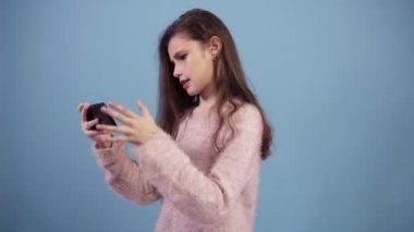 Uzun saçlı beyaz genç kız gülümseyerek ve heyecanla stüdyoda mavi arka plan üzerinde akıllı telefon video oyunu oynarken. Genç kız video oyunu oynarken, gesturing, gerçekten dahil