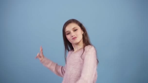 Menina de cabelos longos muito nova em um fundo azul sorrindo e brincando e dançando brincando. Vestindo camisola rosa brilhante, olhando para a câmera. Movimento lento — Vídeo de Stock