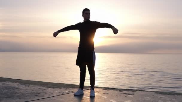 Silhouette di giovane uomo alto in pantaloncini che si scaldano sul pavimento in legno vicino al mare. Allungando le braccia fino all'alba del mattino. Scaldarsi, allenarsi all'aria aperta — Video Stock