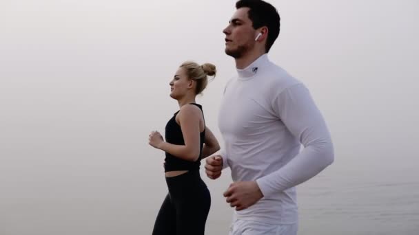 Running paar lopers man en vrouw op het strand opleiding buiten in de buurt van de zee in mistig weer. Jonge knappe man in het wit woth draadloze koptelefoon en meisje joggen samen op zee. Slow Motion — Stockvideo