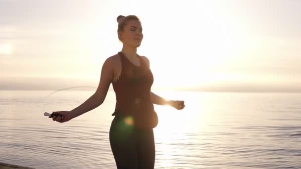 Портрет здоровой молодой женщины с скакалкой на берегу моря. В солнечный день женщины занимаются спортом на свежем воздухе. Утренний мягкий солнечный свет на фоне — стоковое видео