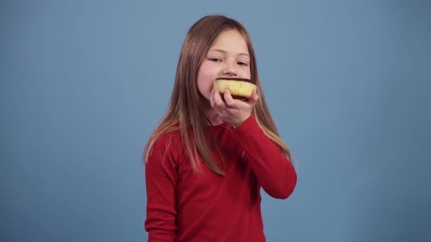 Το κοριτσάκι με το κόκκινο πουλόβερ τρώει ένα ντόνατ σοκολάτας σε χρωματιστό φόντο. Απολαμβάνω τη γεύση. Παιδί 7-10 χρονών, ωραία μαλλιά. Πυροβολισμός στο στούντιο. Έννοια του φαγητού — Αρχείο Βίντεο