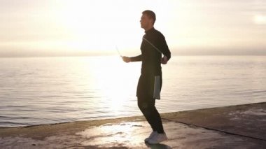 Deniz kenarında ip atlama ile egzersiz beyaz kablosuz kulaklıkile uzun boylu, kaslı genç adam tam uzunlukta. Genç adam sporla uğraşıyor. Yavaş çekim, güneş ışığı