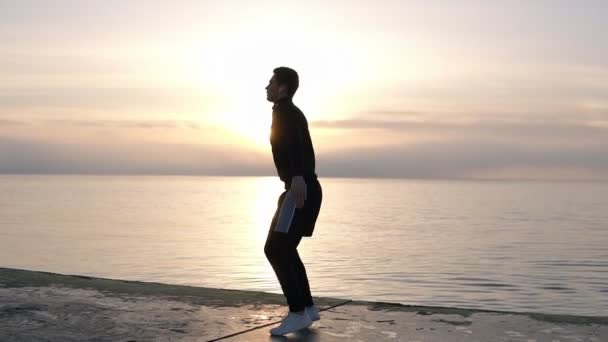 Привлекательный, спортивный высокий мужчина в шортах греется на деревянном полу у моря. Делать приседания с прыжками вверх, упражнения ног. Разминка, упражнения на открытом воздухе. Вид сбоку — стоковое видео