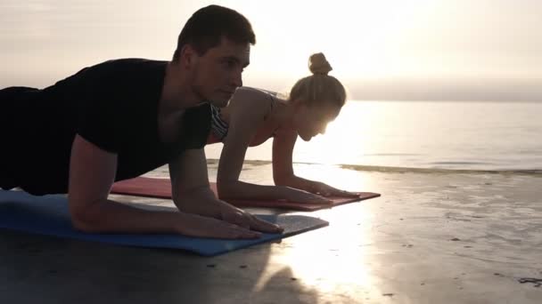 Skoncentrowany kaukaski mężczyzna i blond dziewczyna trzyma pozycję deski na maty do jogi podczas treningu na zewnątrz, w pobliżu morza. Piękne miękkie wschód słońca na tle — Wideo stockowe