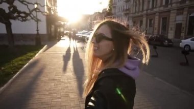 Kahverengi, siyah deri ceket ve siyah güneş gözlüğü uzun saçlı kız yürür hatta güneş ışığı dolu şehir sokak üzerinden çalıştırın. Kız adamların elini çekiyor. Bir randevuda kadın ve erkek, eğleniyor
