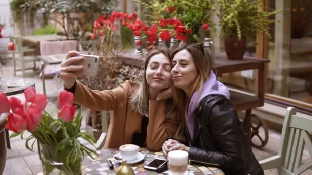 Dos chicas están haciendo selfie. Cambian las expresiones faciales, una chica besando a su amiga a la mejilla. Hacen varias tomas diferentes. Las chicas están sentadas en el café. — Vídeos de Stock