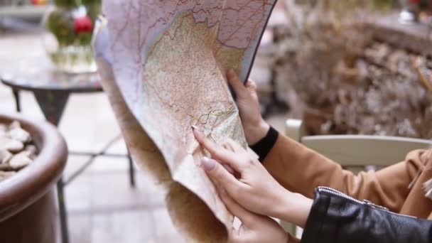 Κλείσιμο του χάρτη-δύο όμορφες Καυκάσιες κοπέλες που κάθονται στο υπαίθριο καφέ του δρόμου με απλωμένο χάρτη. Σχεδιάζουν το ταξίδι τους ή περπατούν στη νέα πόλη. Ένα κορίτσι που σημαδεύει το χάρτη με ένα δάχτυλο. Θολή — Αρχείο Βίντεο