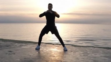 Kaslı, şortlu sportif uzun boylu adam denize yakın ahşap zeminde ısınıyor. Egzersizler bacaklar, yanlara yaslanarak germe. Isın, açık havada egzersiz. Ön görünüm