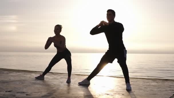 Jovem determinou esportista caucasiano e sua parceira com pernas esticadas antes de correr todos os dias na praia. Manhã sunligth no fundo — Vídeo de Stock