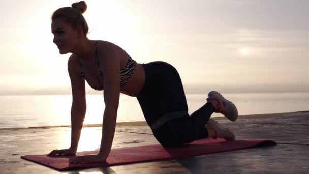 Blízko k záchvatu atletické běločké ženy v sportovní přísaze pomocí elastického pásu s roztažným nohama a se vším tělem u moře na ranní sluneční erupci. Zpomaleně — Stock video