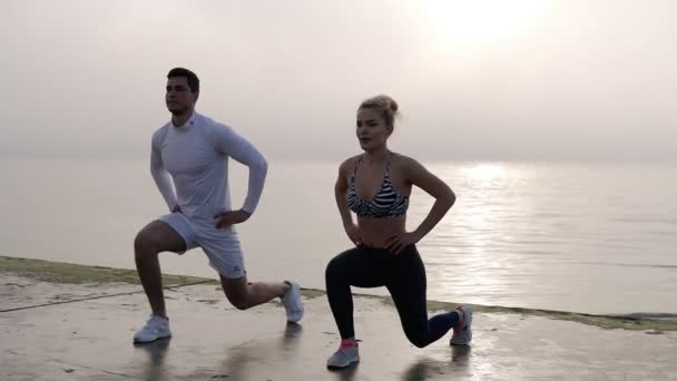 健身,锻炼,户外体育活动概念。一大早,专业的健身人士在海边锻炼。同步向前冲。穿着白色衣服和女人打底裤的男人 — 图库视频影像