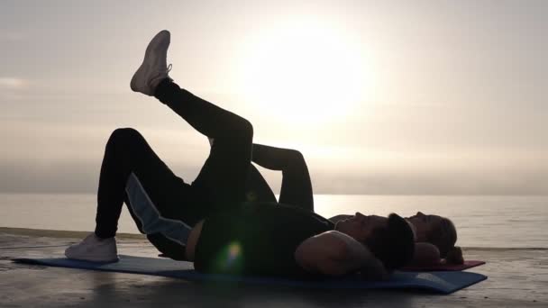 아침 바닷가에서 함께 운동하는 젊은 백인 부부에게 적합합니다. 맞는 여자와 남자 하 고 프레스 업 그들의 엉덩이를 들어 올려. 부드러운 햇빛 배경입니다. 외부에서 훈련하는 운동 선수 — 비디오
