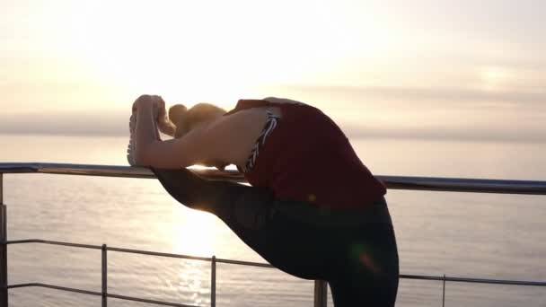 Disparo en cámara lenta de una joven deportista concentrada estirando las piernas en un embarcadero de madera con valla metálica. Hermosa chica rubia entrenando en la playa. Concepto de deporte, fitness — Vídeos de Stock