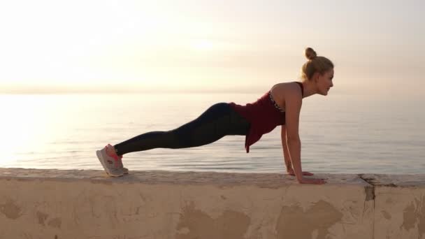 Mignon ajustement fille caucasienne en leggings de sport faire de l'exercice de planche sur la plage au coucher du soleil sur parapet. Fitness, entraînement et mode de vie sain concept. Ciel bleu le matin — Video