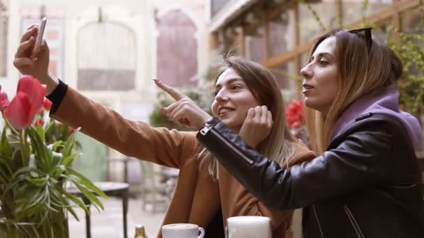 Girlfriends nem selfie a mobil telefon kamera ülve egy kávézóban. Két vonzó lányok képeket a kamera a smartphone és hülyéskedik, mutatva a képernyőn — Stock videók