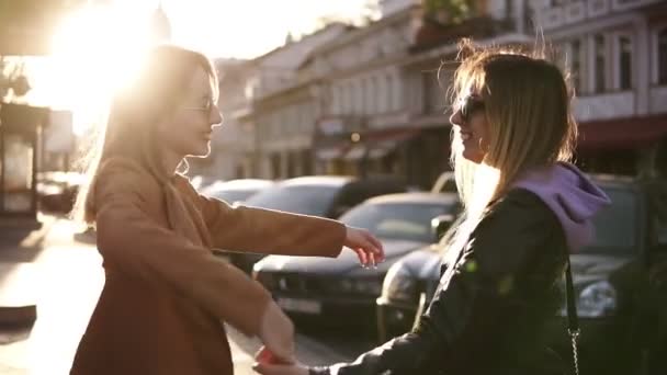 Två lyckliga unga flickor gå av staden tillsammans, krama varandra. Honor omfamna, skratta och upphetsad. Kvinna vänskap, promenad i staden utomhus. Stadsutsikt, solljus på bakgrunden — Stockvideo