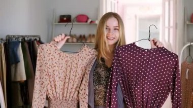 Sarışın uzun saçlı kız eğlenceli ne satın almak için karar veren bir ayna önünde karşılaştırarak askıları iki trendy bluz arasında seçim. Kıyafet butik gülümseyen, neşeli kız