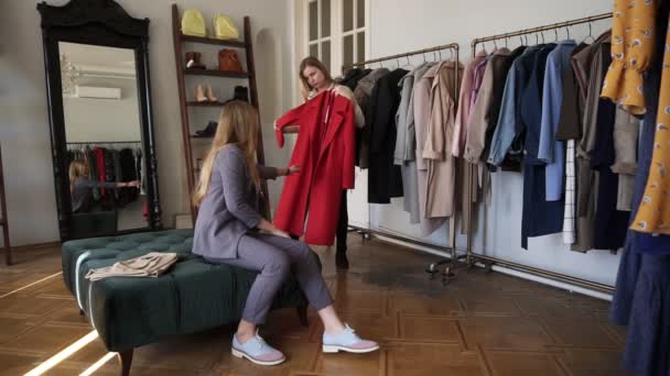 Две девушки или сёстры-близнецы ходят по магазинам вместе: длинноволосая молодая блондинка примеряет новое красное пальто, стоя рядом со стойками с разнообразными нарядами на вешалках. Обсудите ее выбор с сестрой. — стоковое видео