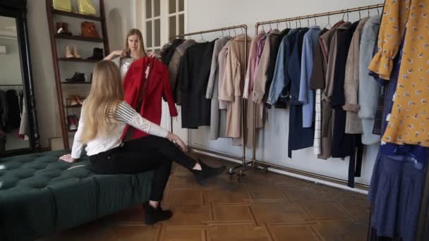 Två flickor eller tvillingsystrar shopping tillsammans: långhårig ung blond kvinna försöker en ny grå och röda rockar stående nära rack med olika outfits på galgar. Diskutera hennes val — Stockvideo