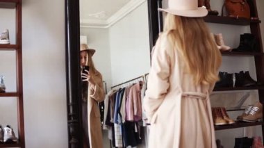 Çekici sarışın kız bej ceket ve trendy şapka ayna önünde duran, onun akıllı telefon ile bakmak bir fotoğraf yapma. Arkadan görüntüler