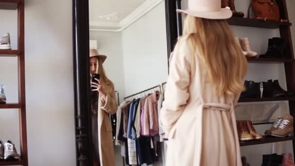 Привлекательная блондинка, стоящая перед зеркалом в бежевом пальто и модной шляпе, фотографирует себя со своим смартфоном. Видеозапись с задней стороны — стоковое видео