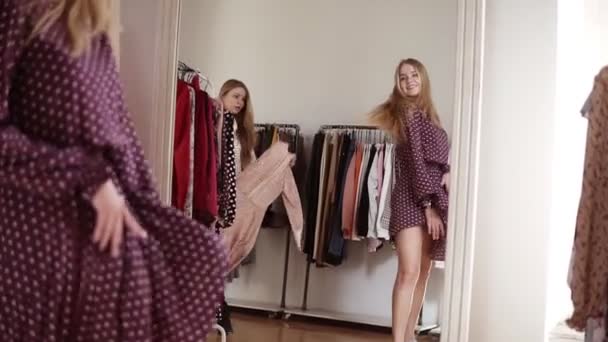 Twee blonde tweeling zusters zijn samen aan het winkelen. Men probeert de trendy jurk aan de voorkant van de spiegel, zuster biedt aan haar een andere jurk te proberen. Staande in de voorste spiegel in de buurt van het rek met kleding op hangers — Stockvideo